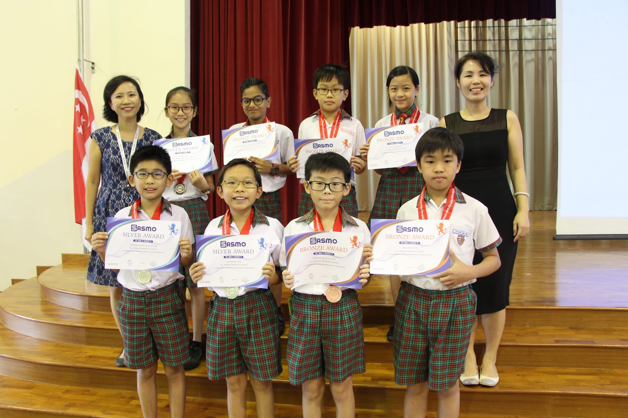 Eight students in school uniforms hold SASMO Silver and Bronze Award certificates, flanked by two adult women on a stage.